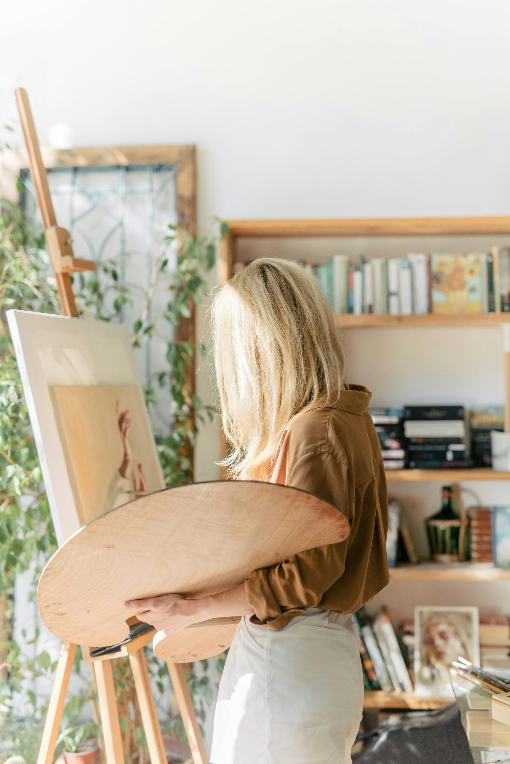 A woman artist with blonde hair paints on canvas in a cozy, sunlit studio filled with books and art supplies.